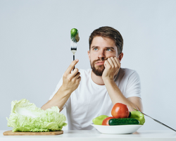 A man, surrounded by different vegetables, looking at a fork with a cucumber (Success Through Failure episode 341: Finally, The One Diet To Rule Them All: Guaranteed Results)